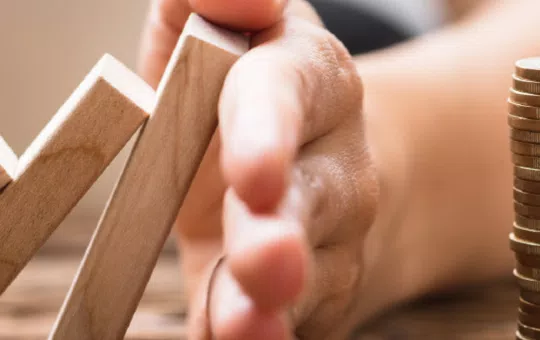 A hand is stopping a set of wooden blocks arranged in ascending order, preventing them from toppling. In the background, there is a stack of coins on a wooden surface.