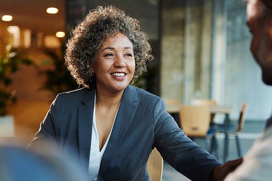 A smiling woman in a gray suit engages in conversation, reaching out to shake hands with someone off-screen, in a modern office environment with soft lighting and indoor plants.