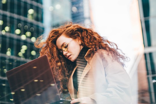 A young woman with curly red hair types on a laptop, focused and deep in thought, while surrounded by tall city buildings and a blurred background of urban activity.