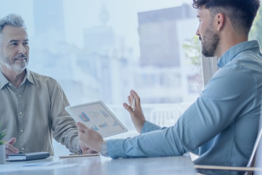 Two men are engaged in a discussion at a table, one gesturing with an electronic tablet displaying graphs, while the other listens attentively in a modern office setting with large windows.