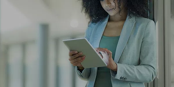 A woman in a light gray suit is intently using a tablet while leaning against a wall in a modern, well-lit environment with blurred backgrounds suggesting an office setting.