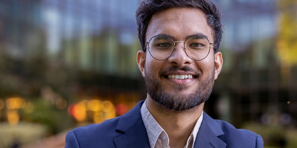 A man with glasses and a beard smiles confidently while wearing a blazer. He stands outdoors in front of a building with large windows, surrounded by greenery and soft, blurred lights.