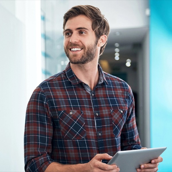 A man wearing a plaid shirt is holding a tablet and smiling. He stands in a modern, well-lit environment with blue accents and blurred background elements.