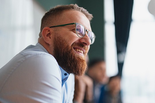 A man with a beard and glasses smiles while seated, engaged in conversation with others in a bright, modern indoor setting. Soft lighting enhances the warm atmosphere.