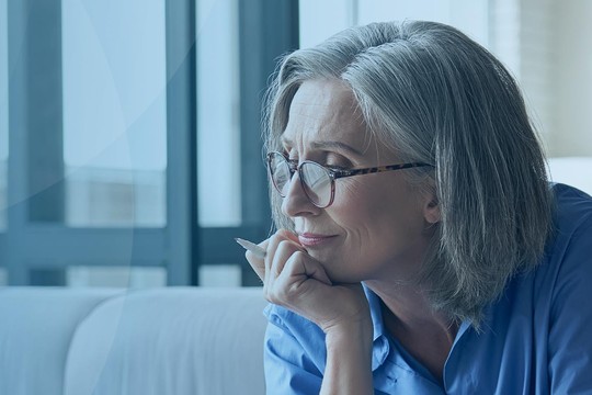 A woman with gray hair and glasses rests her chin on her hand, gazing thoughtfully. She is seated on a light-colored sofa, with bright natural light coming through large windows nearby.