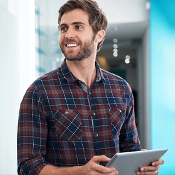 A man holds a tablet while smiling, standing in a brightly lit hallway with modern decor and a blue accent wall. He appears engaged and approachable.