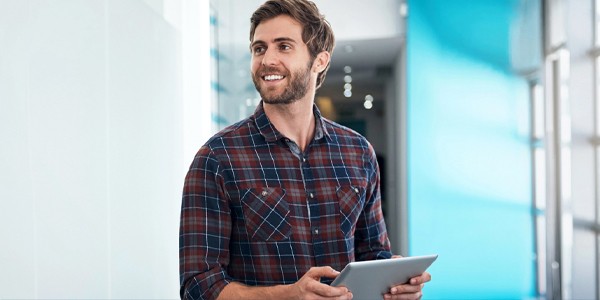 A man holds a tablet while smiling, standing in a brightly lit hallway with modern decor and a blue accent wall. He appears engaged and approachable.