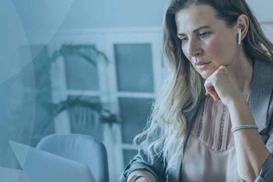 A woman sits at a desk, focused on her laptop, resting her chin on her hand. The background features a light, modern workspace with plants and framed art.