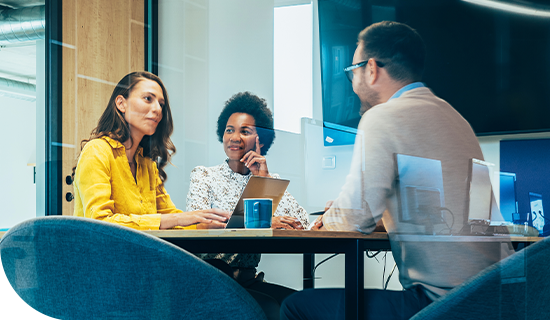 Three professionals sit around a table, engaged in discussion. One woman in a yellow sweater speaks, while another woman listens thoughtfully. A man in glasses contributes, in a modern office setting.