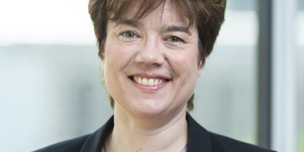 A smiling woman with short, dark hair wears a black blazer over a red top, standing indoors with a soft, blurred background of light windows.
