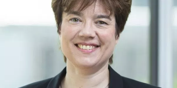 A smiling woman with short, dark hair wears a black blazer over a red top, standing indoors with a soft, blurred background of light windows.