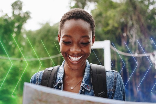 A woman smiles while holding a map, exploring her surroundings. She wears a denim jacket and has a backpack, with lush greenery in the background, suggesting an outdoor adventure.