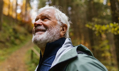 An elderly man with a white beard smiles while looking upward. He is wearing a jacket in a wooded area with soft, diffused light filtering through the trees. A path is visible behind him.