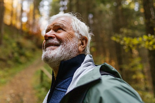 An elderly man with a white beard smiles while looking upward. He is wearing a jacket in a wooded area with soft, diffused light filtering through the trees. A path is visible behind him.