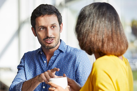 A man in a blue shirt gestures animatedly while speaking to a woman in a yellow top, both engaged in conversation in a brightly lit, modern café setting.
