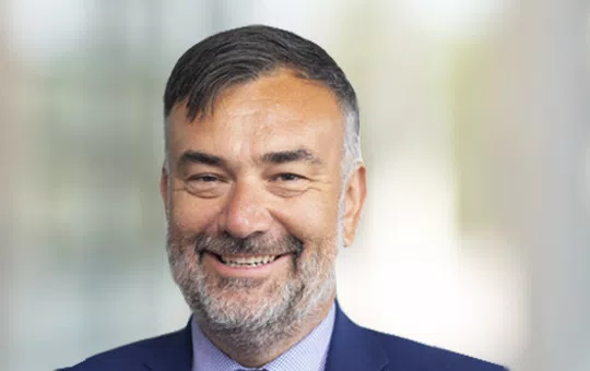 A man with a short beard and neatly styled hair smiles confidently. He wears a dark suit over a light shirt, set against a blurred, indoor background.