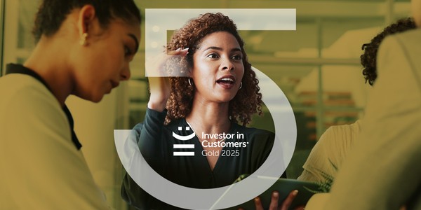 A young man smiles while engaged in conversation with colleagues in a modern office setting. The context includes a bright space with casual seating and a glass wall. The text reads: "Investor in Customers® Gold 2024."