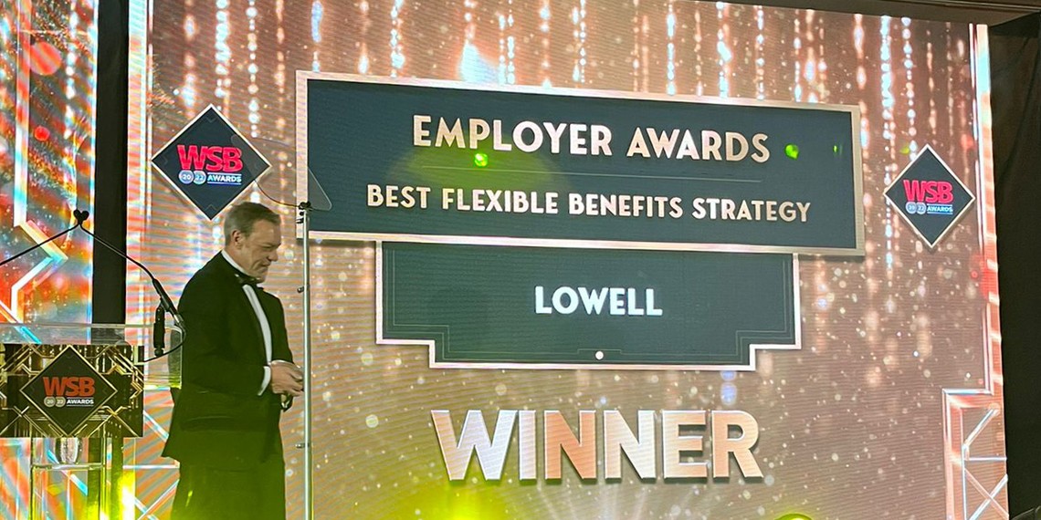 A man in formal attire stands beside a brightly lit awards stage. A prominent display announces "EMPLOYER AWARDS," "BEST FLEXIBLE BENEFITS STRATEGY," "LOWELL," and "WINNER."