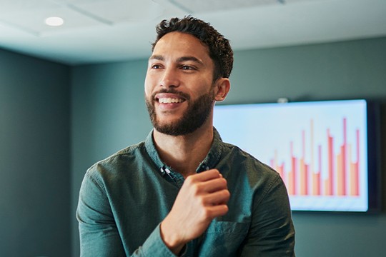 A man with a beard smiles while gesturing, seated in a modern office with a large screen displaying bar graphs in the background, indicating a business meeting or presentation setting.