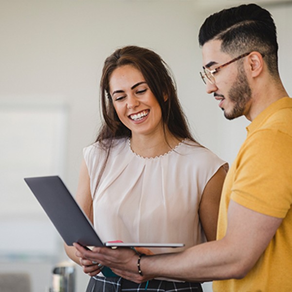A woman and a man smile while discussing a laptop in a modern office setting. The woman points at the screen, engaged in conversation, with a whiteboard and other people visible in the background.