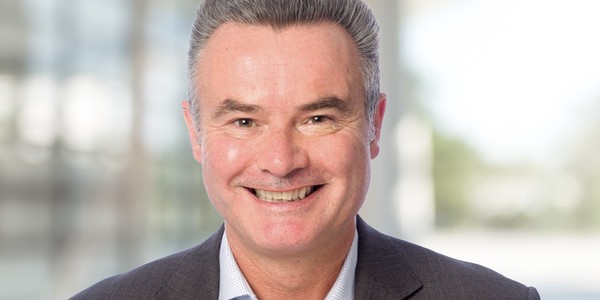 A middle-aged man smiles broadly, wearing a dark blazer over a light shirt. He stands against a blurred background suggesting a modern indoor setting.