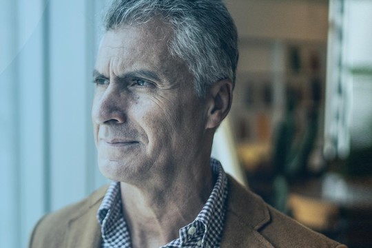 A mature man stands thoughtfully by a large window, gazing out at a modern cityscape, with soft lighting and muted colors in a professional-looking environment.