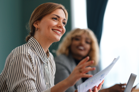 A smiling woman in a striped shirt holds papers, engaging in conversation. A blurred figure in the background shows interest. The setting is an office with natural light and green walls.