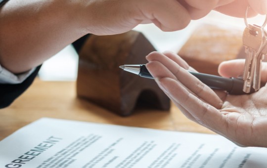 A hand holds house keys, transferring them to another person’s outstretched palm. A signed agreement lies on a wooden table, suggesting a real estate transaction or rental arrangement.
