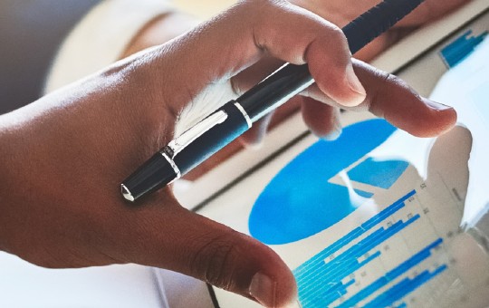 A hand holds a pen while interacting with a tablet displaying blue graphs and data visualizations. The setting suggests a professional or analytical environment, possibly for business or research.