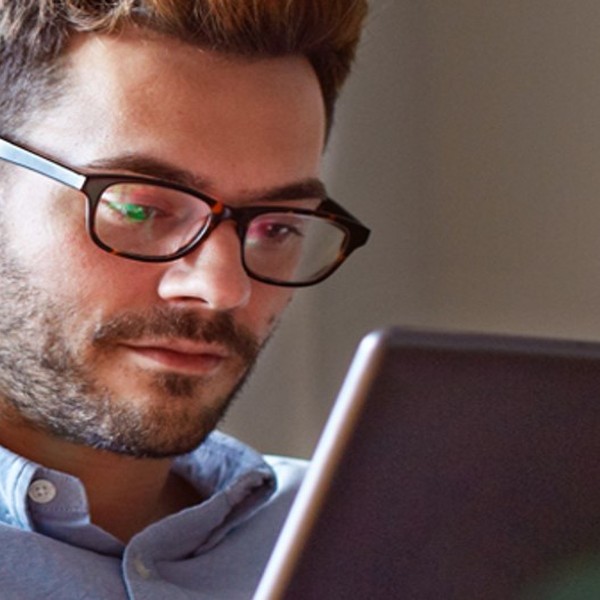 A man with glasses intently uses a tablet while seated in a well-lit, casual indoor environment, showing focus and engagement in his activity.