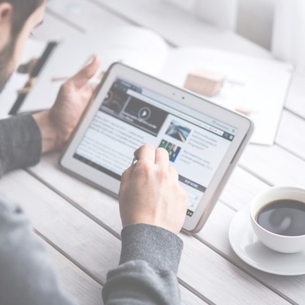 A person sitting at a wooden table interacts with a tablet, reading content while holding a stylus. A cup of coffee sits beside them, and an open magazine is visible nearby.