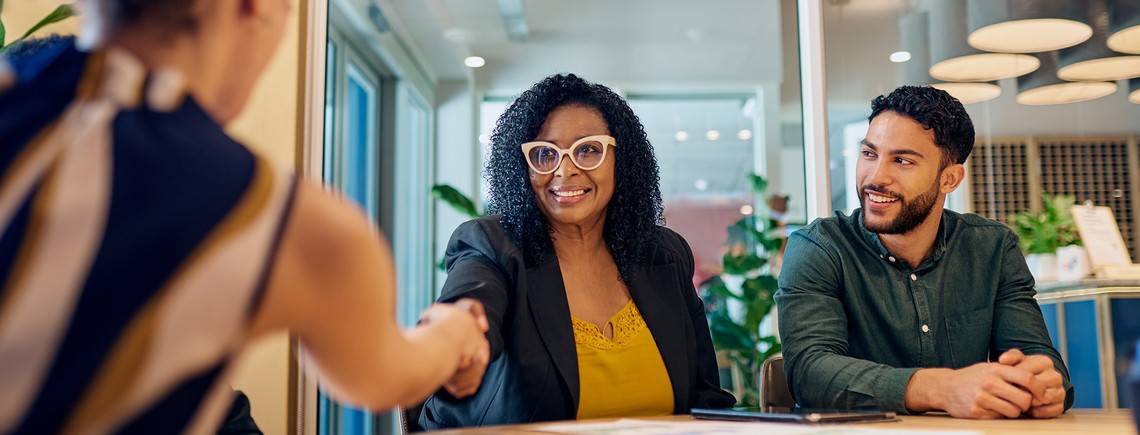 A woman in a black blazer shakes hands with a person offscreen, smiling in a bright, modern office space with green plants and stylish decor. A man sits nearby, observing.